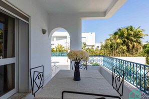 A balcony with a white railing, a vase with white flowers, and a view of palm trees and buildings in the background.