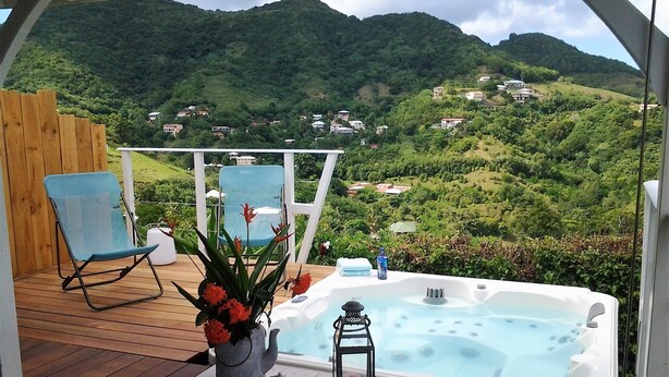 A hot tub on a wooden deck with a view of a green hillside and houses in the distance. - A LOU, A haven of peace with its private jacuzzi for nature lovers ! (Le Diamant)