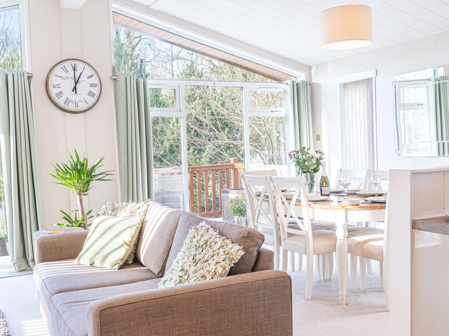 Un salon confortable avec un canapé, une table à manger entourée de chaises, une horloge au mur et de grandes fenêtres avec des rideaux.