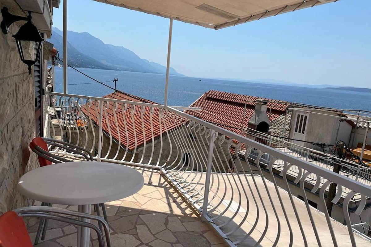 A balcony with a white table and red chairs, overlooking a sea and mountains.