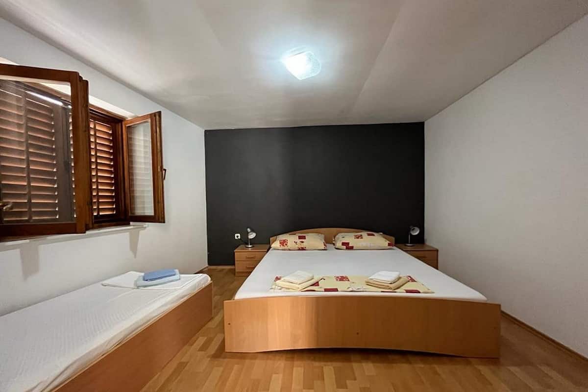 A bedroom with a bed, a bedside table, a nightstand, a window with blinds, and a ceiling light.