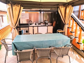 A covered outdoor dining area with a table and chairs, a kitchen in the background, and a staircase with orange balusters. - Sea Marble Villa Apartments (Sozopol)