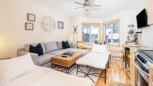 A modern living room with a grey sofa, a wooden coffee table, and a white armchair. There is a ceiling fan, a large window with a view of trees, and a kitchen area with a stove and cabinets.