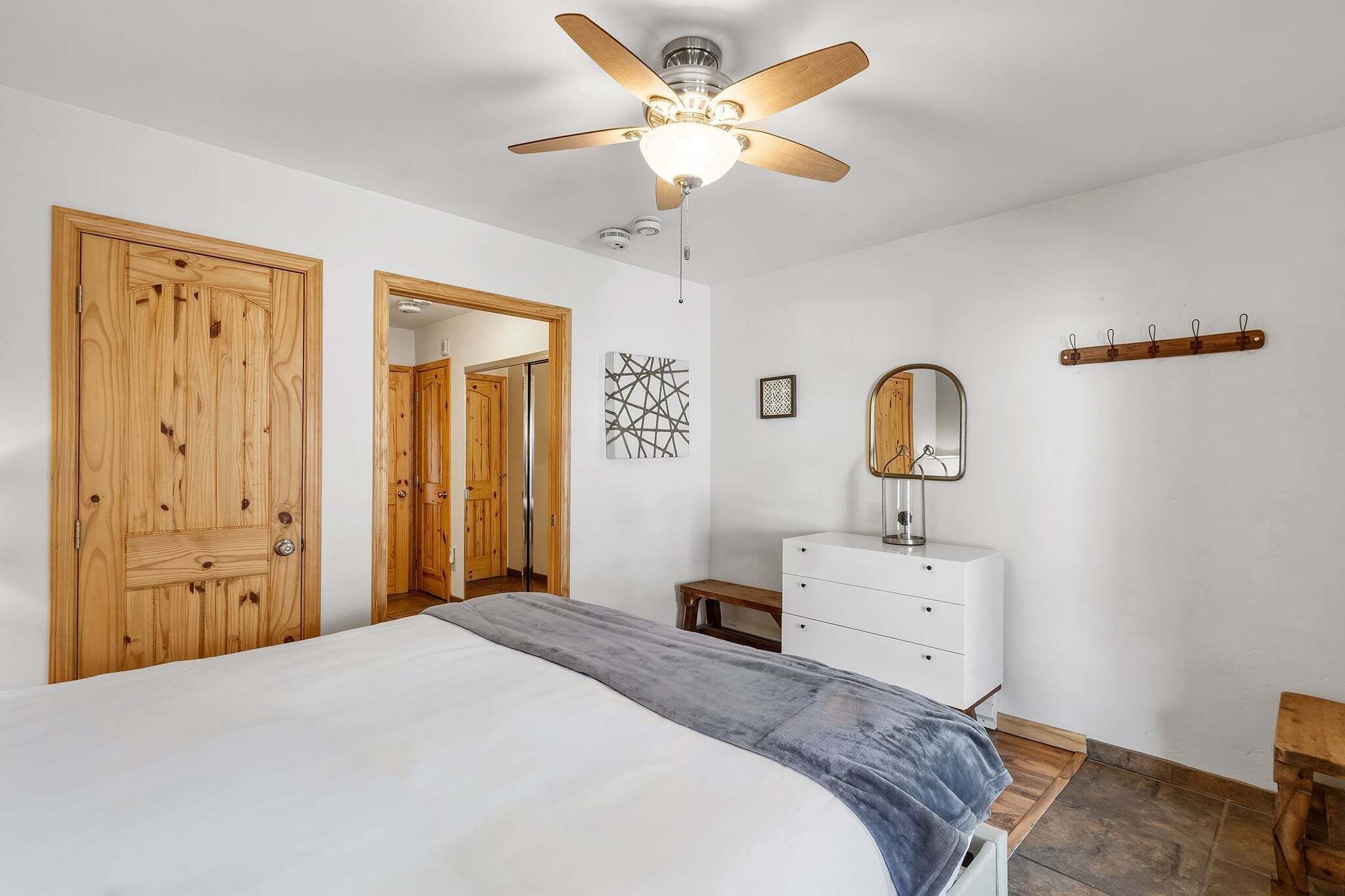 A bedroom with a bed, wooden wardrobe, white dresser, ceiling fan, and a mirror.