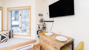 A cozy room with a bed, a dining table set for two, a TV mounted on the wall, and a window with a view of trees.