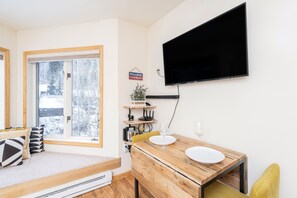 A compact living space with a dining area, a mounted TV, and a window with a view of snow-covered trees. - Modern Comfort River/mtn Views + Hot Tub 1 Bedroom Condo by RedAwning (Telluride)