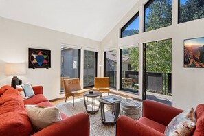 A modern living room with a red sofa, a coffee table, and a fireplace. There are large windows providing a view of the outdoors. - Hidden Gem, 4 Min To Lift 7 Garage, Hot Tub, Lux Kitchen, Big + Bright 4 Bedroom Condo by RedAwning (Telluride)