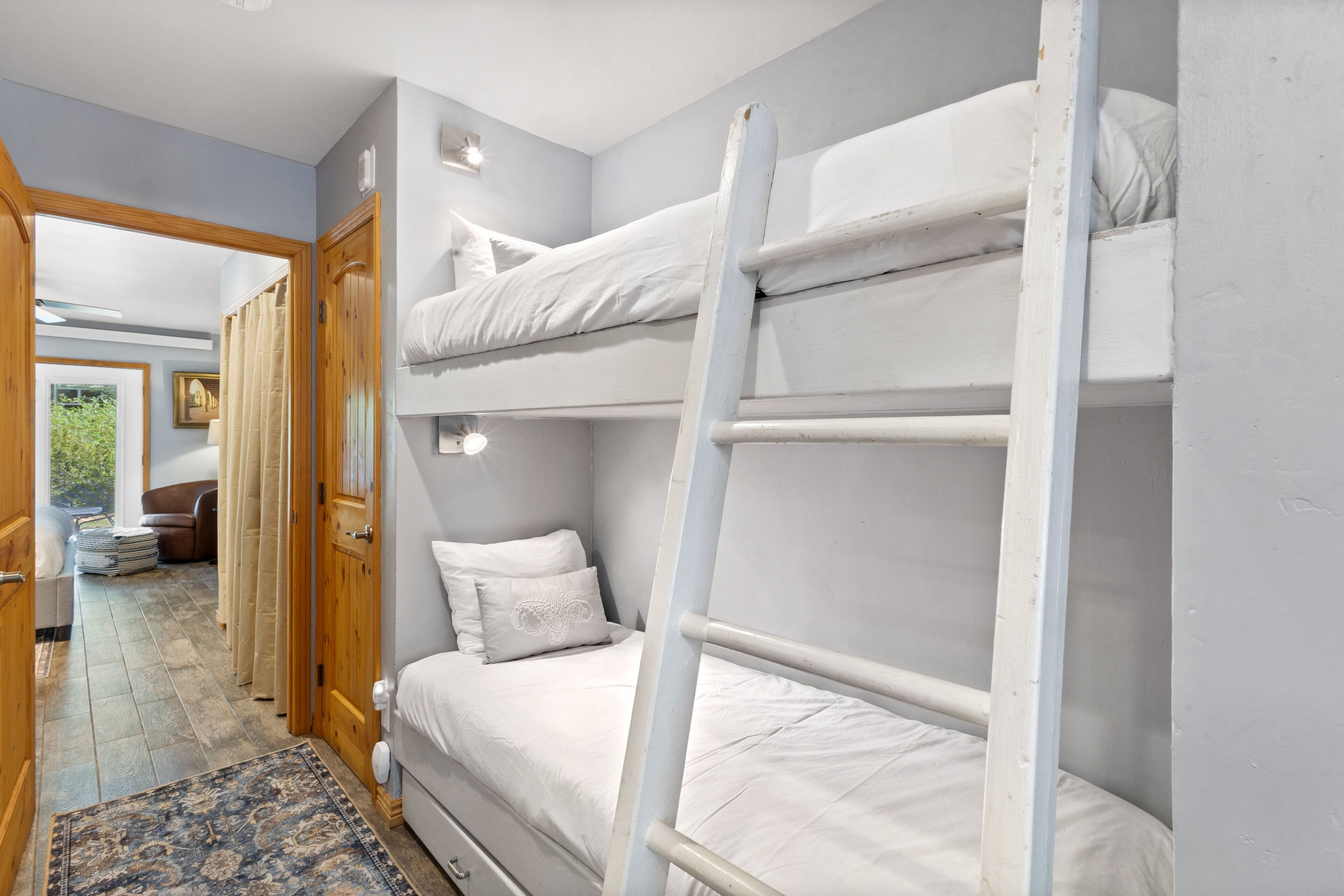 A bunk bed room with a wooden ladder, a door leading to another room, and a patterned rug on the floor.