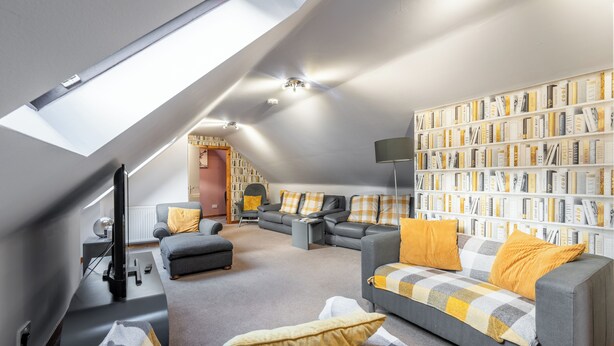 Un moderno soggiorno in stile loft con soffitto spiovente, una libreria a muro e un angolo salotto accogliente con divano e poggiapiedi grigi.