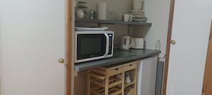 A compact kitchen with a microwave, kettle, and stacked dishes on shelves.