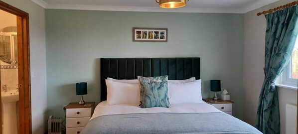 A neatly made bed with a dark headboard, two bedside tables with lamps, a framed picture above the bed, and a window with curtains.