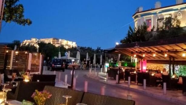 An outdoor dining area with tables set for two, a covered seating area, and a view of a building in the background. - Luxurious Apartment in Acropolis (Athens)