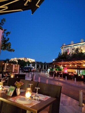 An outdoor dining area with tables set for two, a covered seating area, and a view of a building in the background. - Luxurious Apartment in Acropolis (Athens)