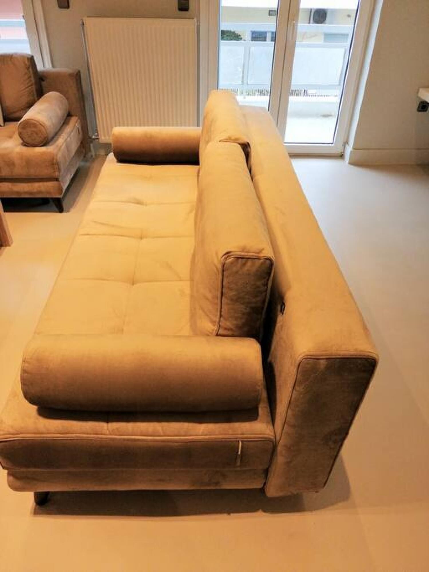 A beige sofa in a room with a large window, a radiator, and a door leading outside.