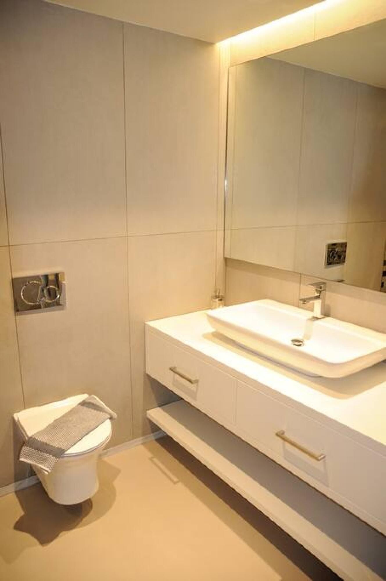 Egy modern fürőszoba, fehér mosdókagylóval, tükörrel és egy WC-vel, melyhez tartozik egy papírtörlő-tartó.