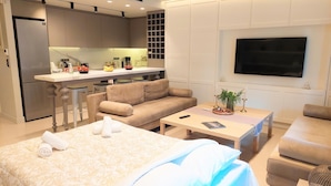 A modern hotel room with a bed, two sofas, a small table, a TV, a kitchen area with a fridge and counter, and a built-in shelf. - Luxurious Apartment in Acropolis (Athens)