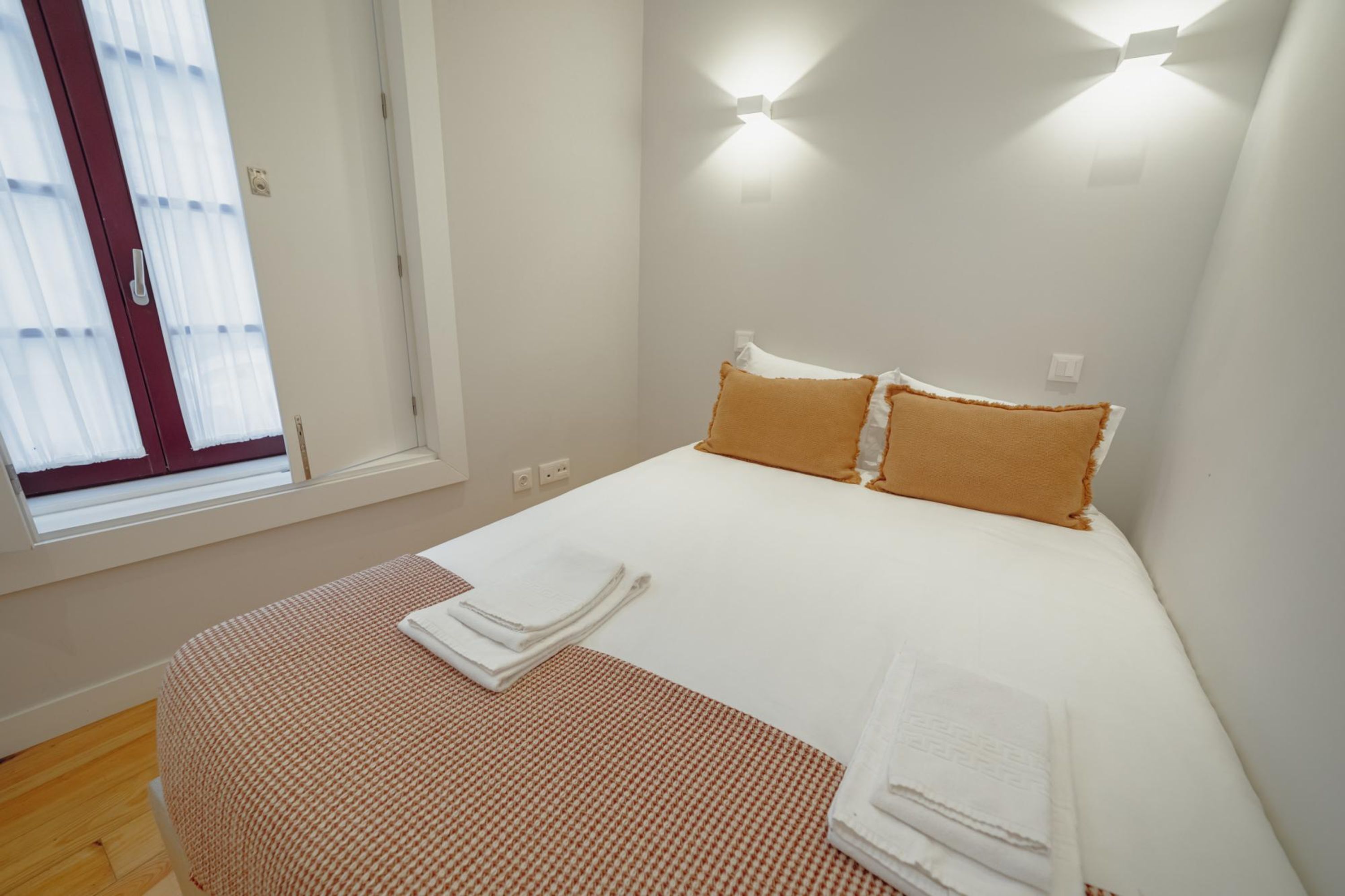 A neatly made bed with two pillows, a window with sheer curtains, and a wall-mounted light fixture.