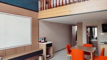 A modern hotel room with a large bed, a desk, and a dining area with orange chairs.