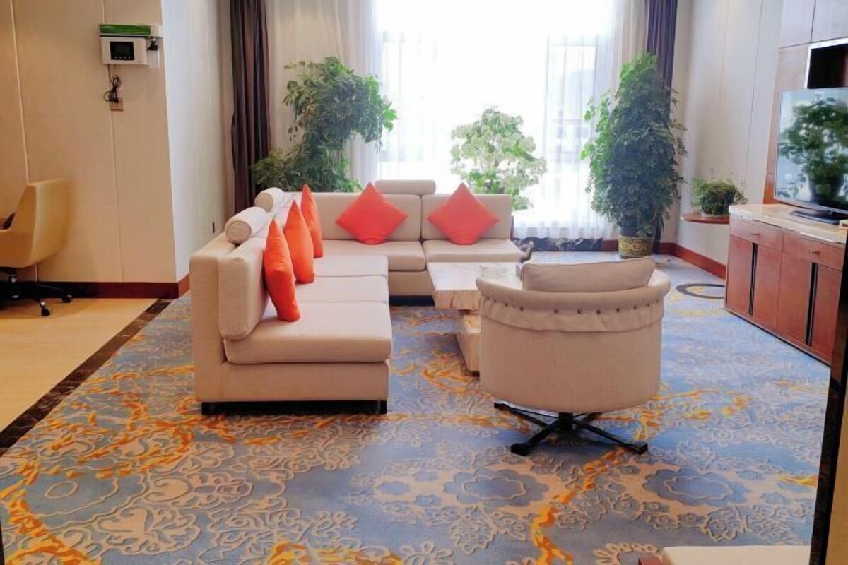 A modern living room with a beige sofa, a white armchair, and a television. There are two potted plants and a large window with curtains.