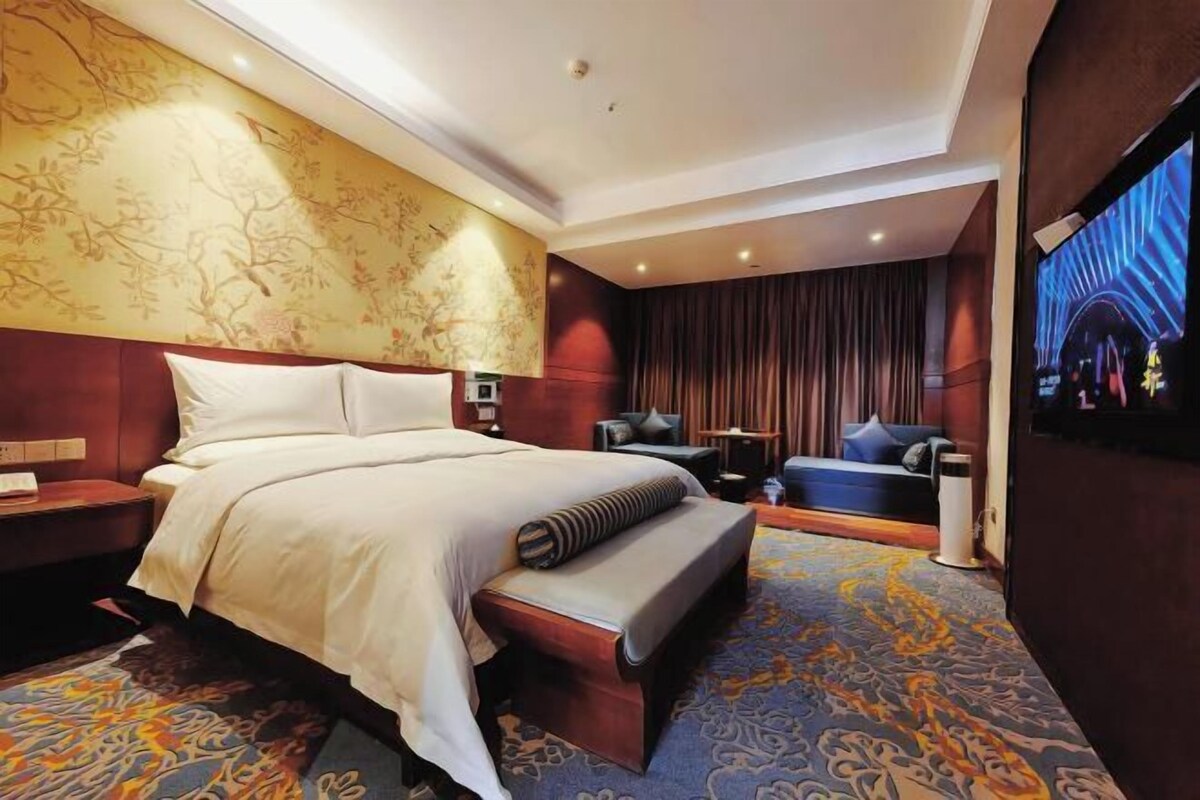 A hotel room with a large bed, a seating area with a sofa and armchairs, a television, and a patterned carpet.