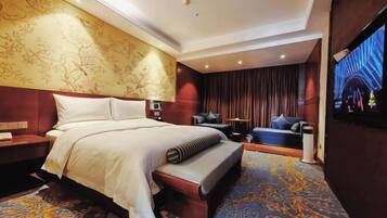 Habitación de hotel con una cama grande, una zona de estar con sofá y sillones, televisión y alfombra estampada.