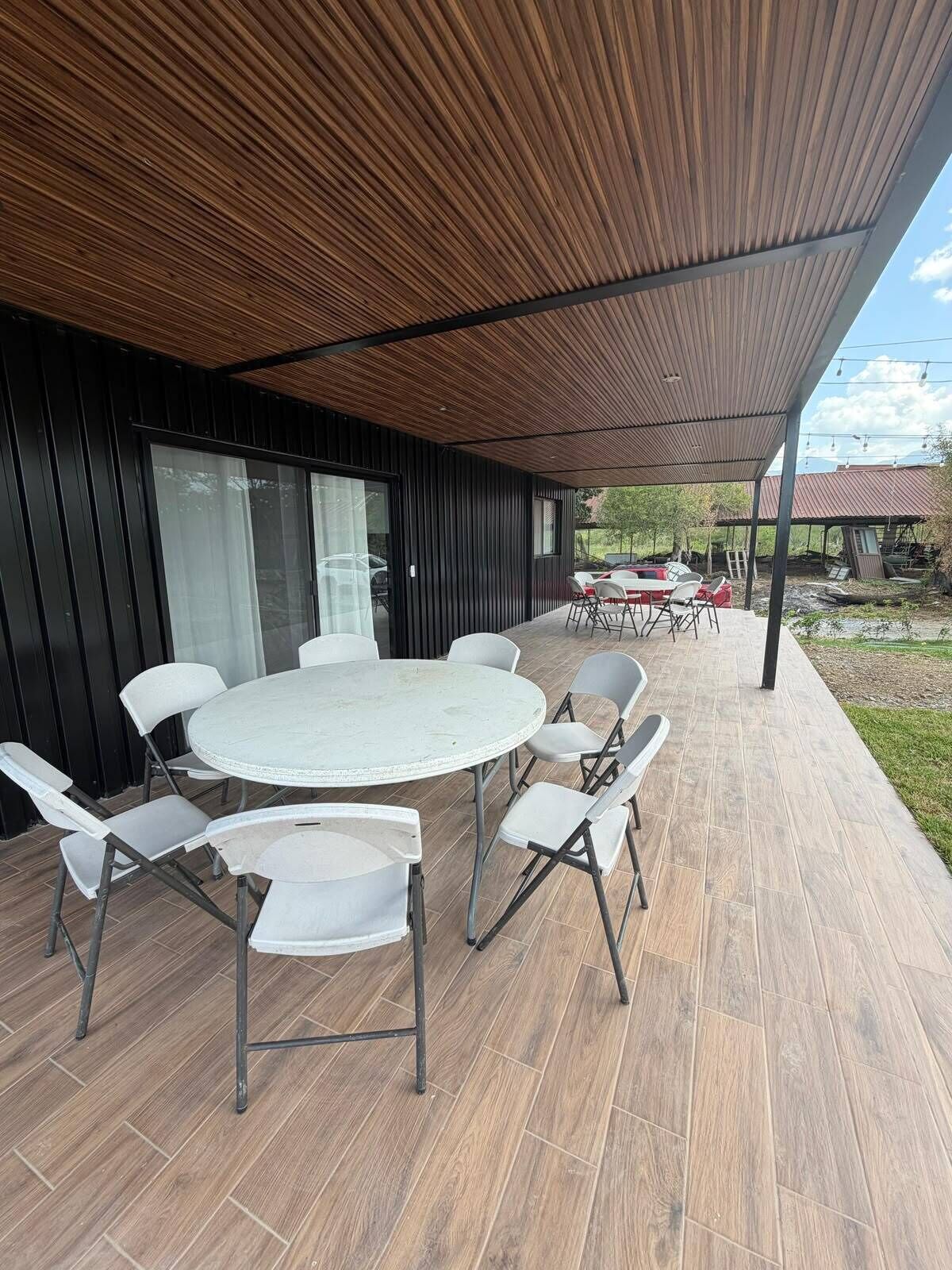 An outdoor patio with a round table and chairs, a covered area with a wooden ceiling, and a view of the surrounding landscape.