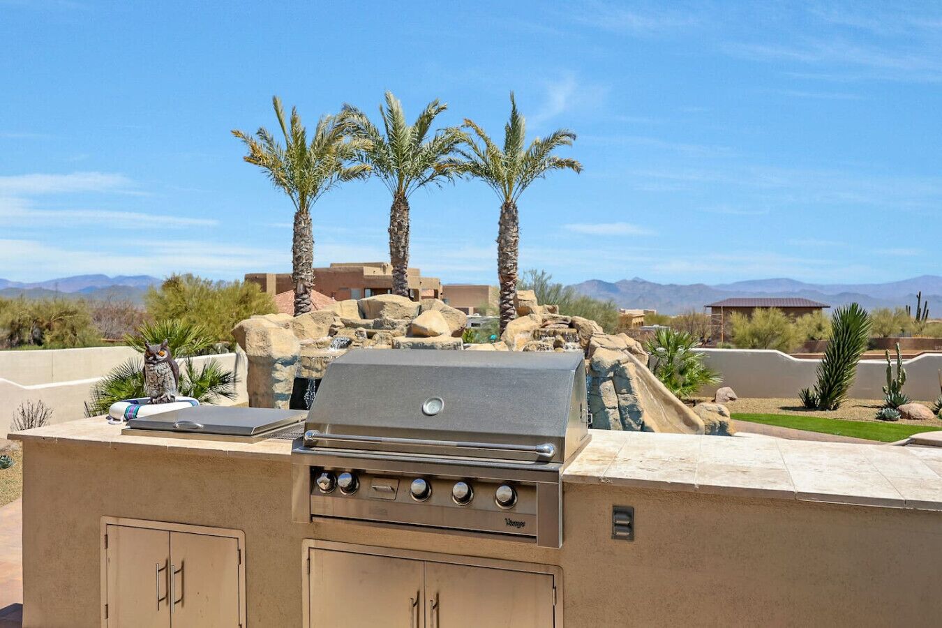An outdoor grill area with a stone wall, palm trees, and a view of mountains.
