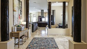 A spacious hallway with a kitchen area, featuring a marble floor, dark wooden columns, and a large abstract painting on the wall.