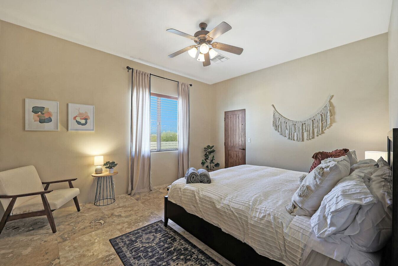 Quarto com cama, cadeira, mesa pequena, ventoinha no teto e janela com cortinas.