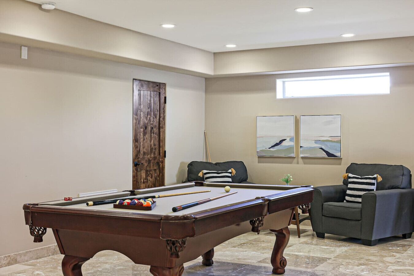Une table de billard dans une pièce où se trouvent deux chaises, une porte et deux tableaux accrochés au mur.