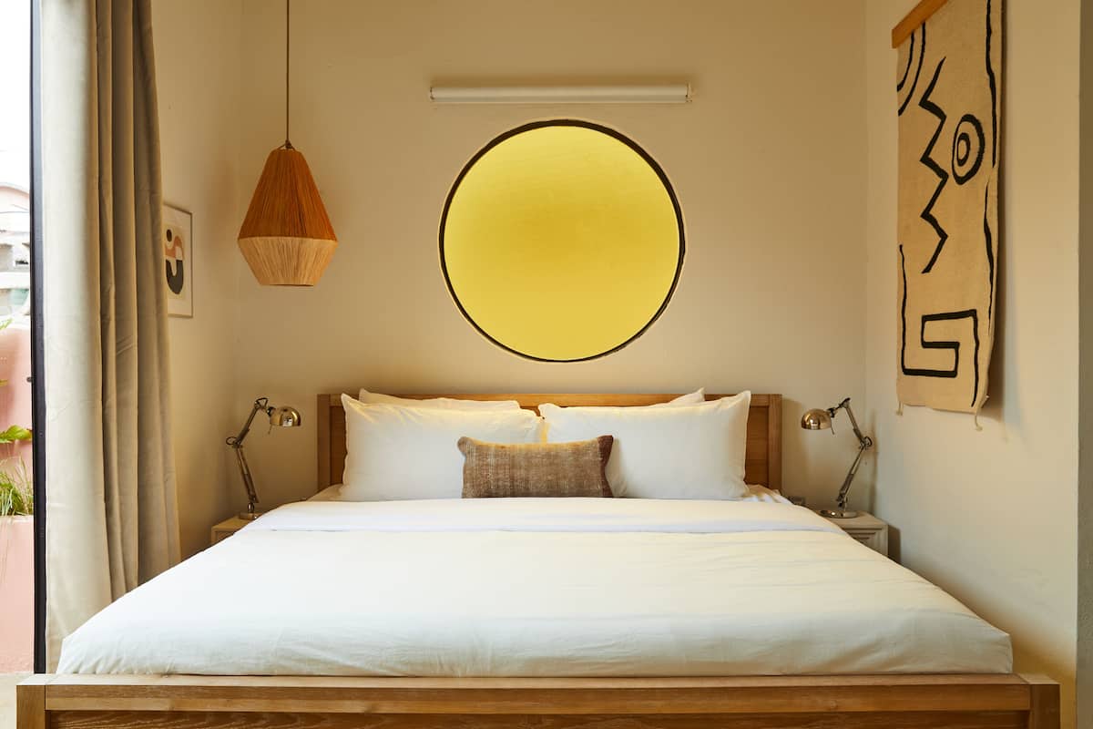 A neatly made bed with white linens and a single decorative pillow, a hanging light fixture, and a wall-mounted circular light.