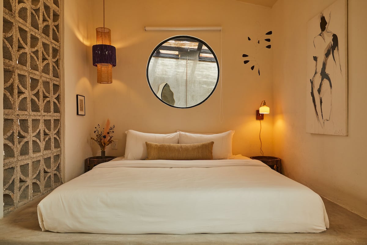 A bedroom with a large bed, a round mirror, a wall-mounted light, and a decorative wall piece.