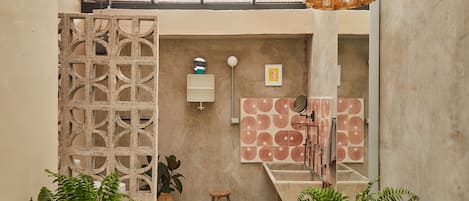 A modern bathroom with a concrete floor, a wooden stool, a decorative wall panel, a ceiling fan, and a hanging light fixture.