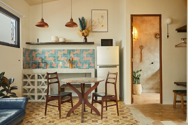Une cuisine compacte avec un réfrigérateur blanc, un four à micro-ondes et un évier. On y trouve une table à manger avec quatre chaises et un canapé bleu. La cuisine est agrémentée d’un crédence décorée et d’un luminaire suspendu.
