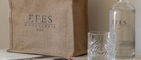 A burlap tote bag with 'EFES HOTELERIE Rome' printed on it, a clear glass bottle with the same branding, and a glass on a table with a magazine.