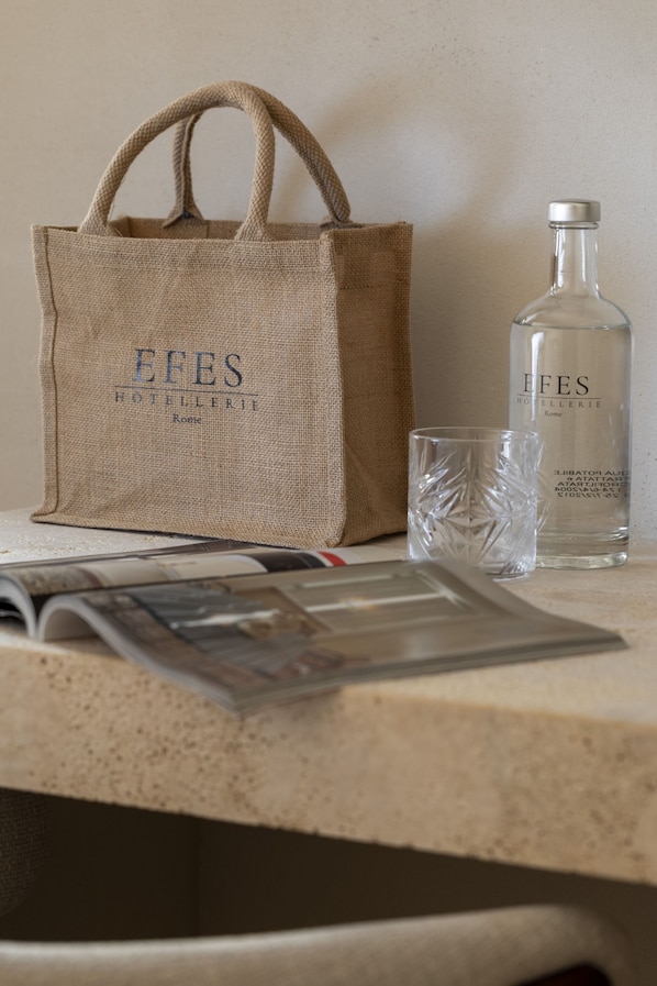 Una bolsa de arpillera con la inscripción 'EFES HOTELERIE Rome', una botella de vidrio transparente con la misma marca y un vaso sobre una mesa con una revista.