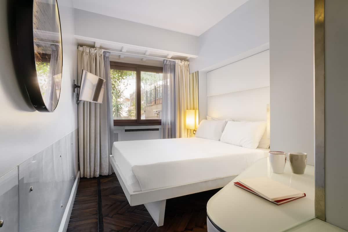 Habitación de hotel con cama, mesita de noche, lámpara y ventana con cortinas.