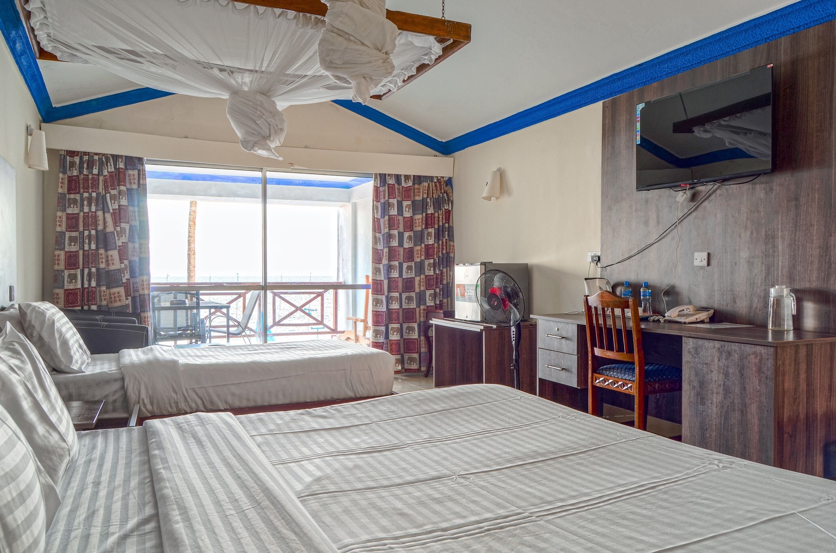 A hotel room with two beds, a desk with a chair, a TV mounted on the wall, and a balcony with a view.