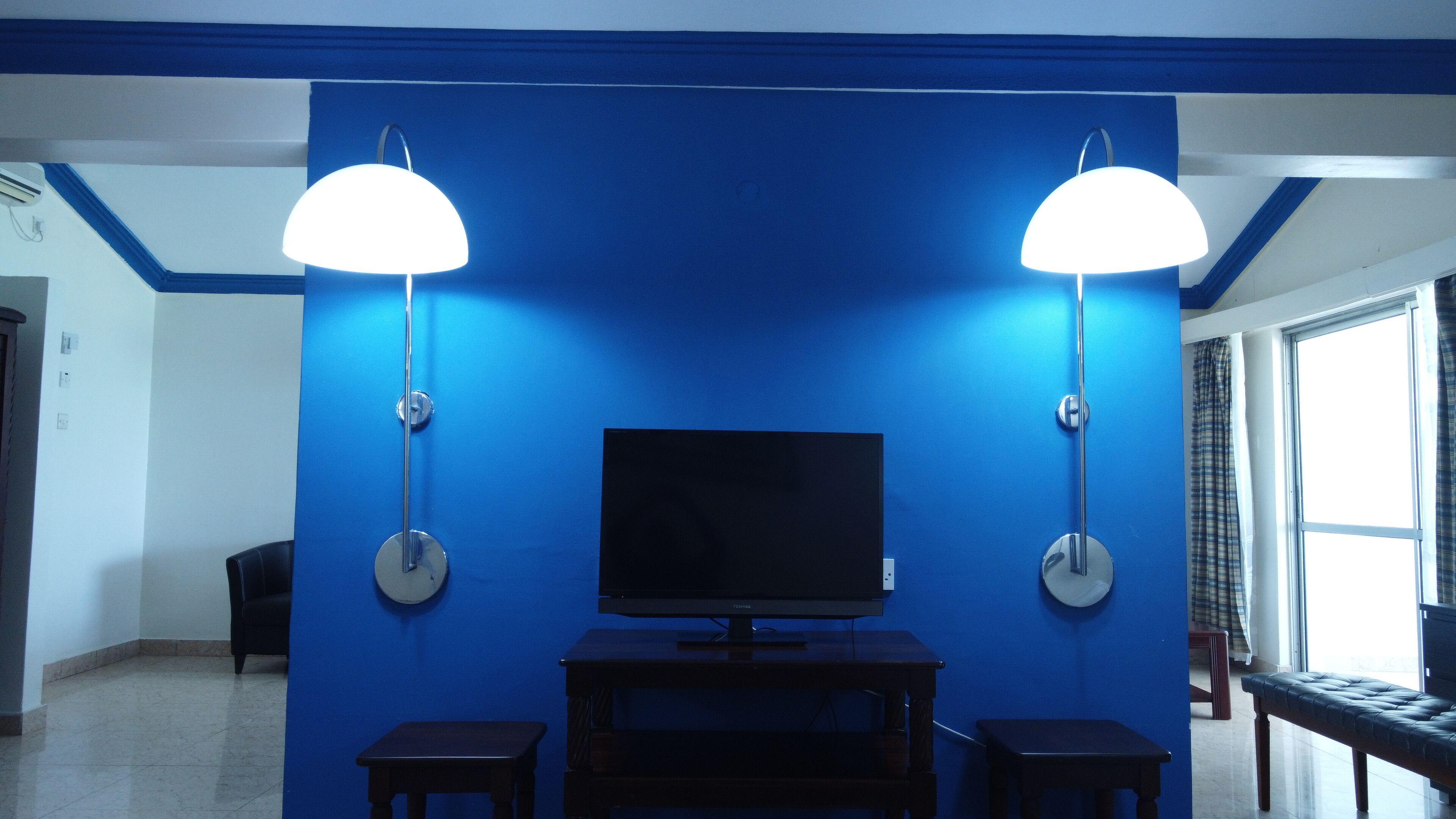 Quarto com parede azul, televisão de tela plana em um suporte, duas luminárias fixadas na parede e um banco.