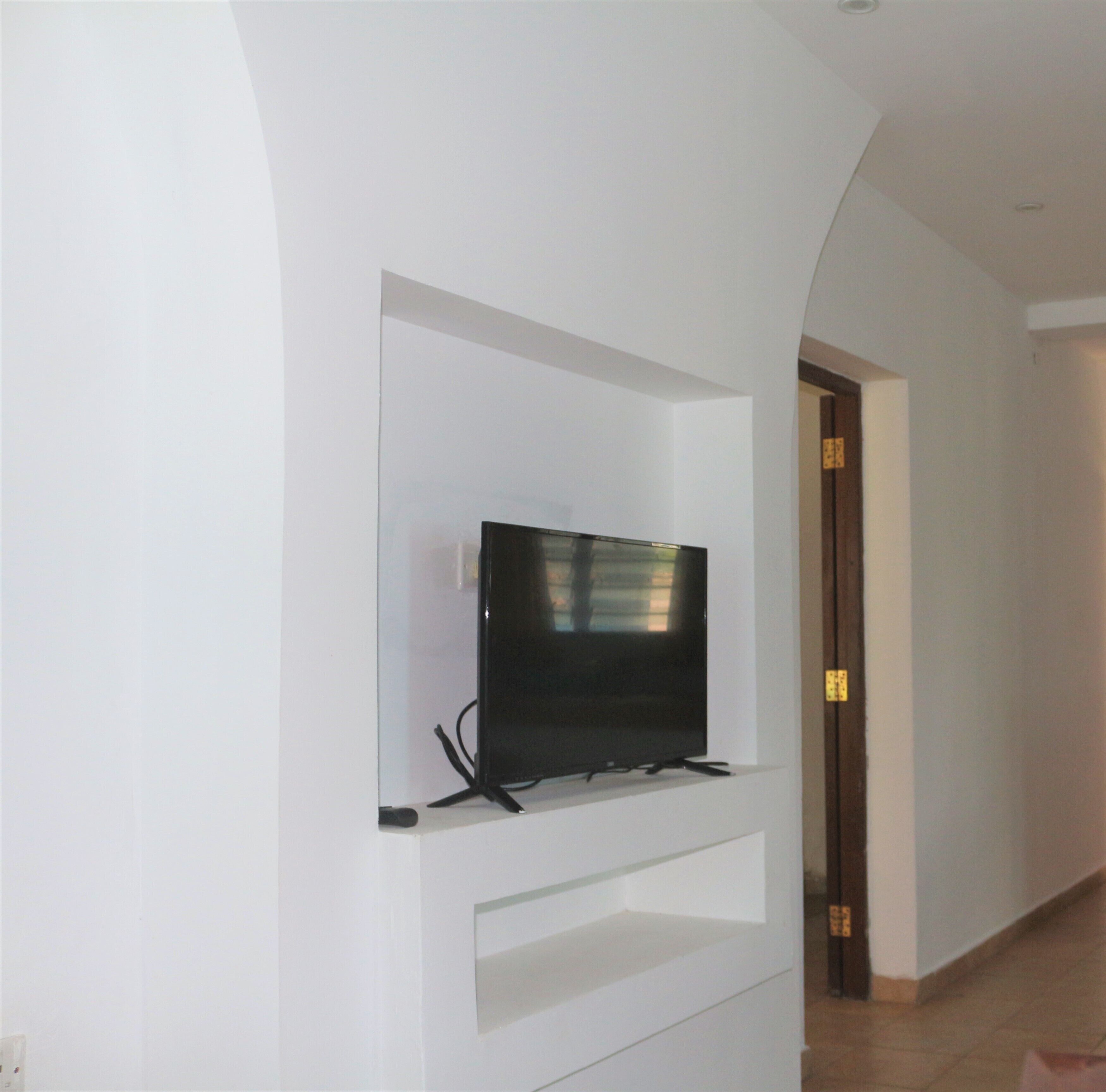 A modern living room with a flat-screen TV mounted on a white wall, a built-in shelf below, and a door leading to another room.