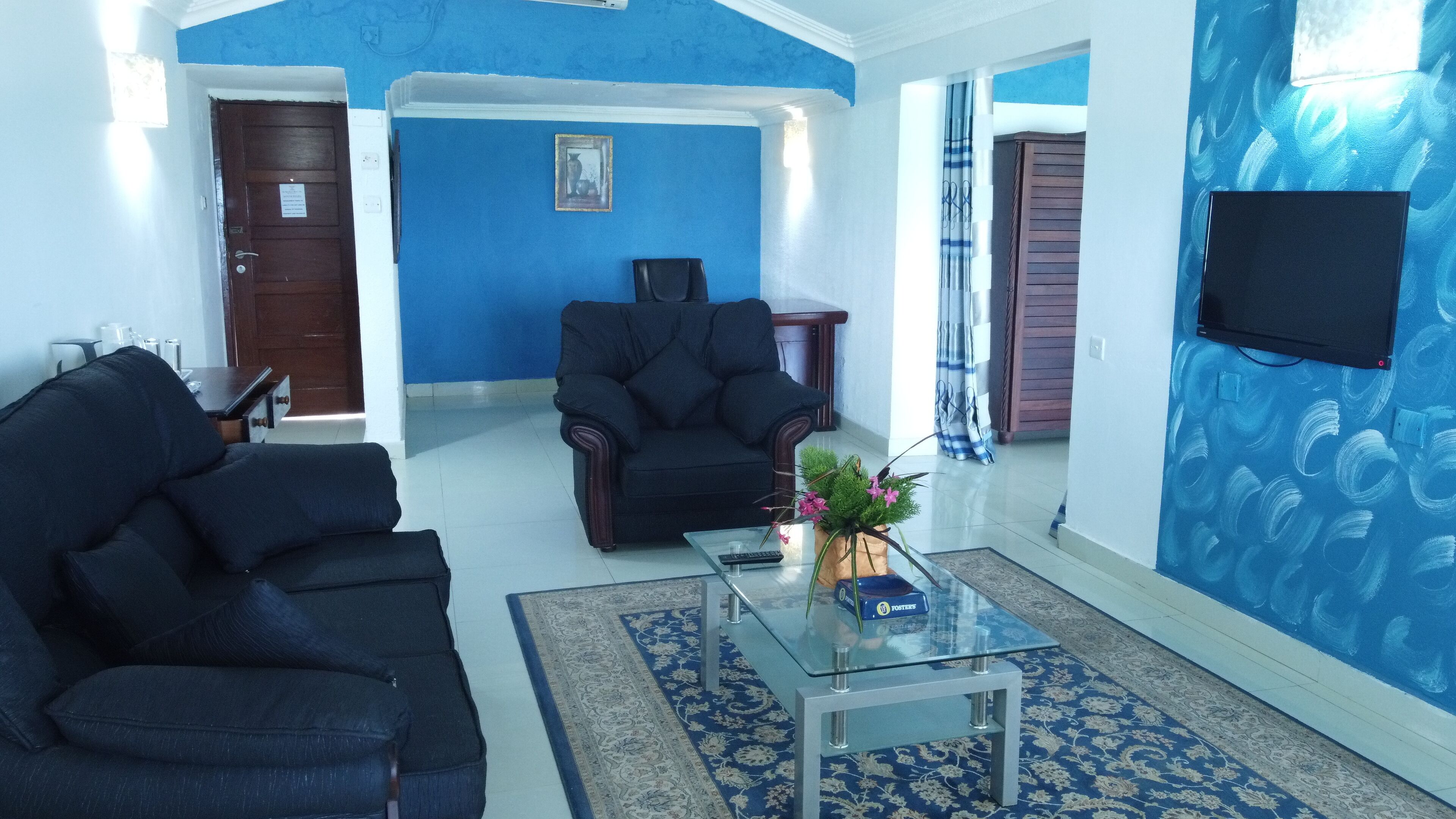 A living room with a blue wall, a black sofa, a glass coffee table with a vase, and a TV mounted on the wall.