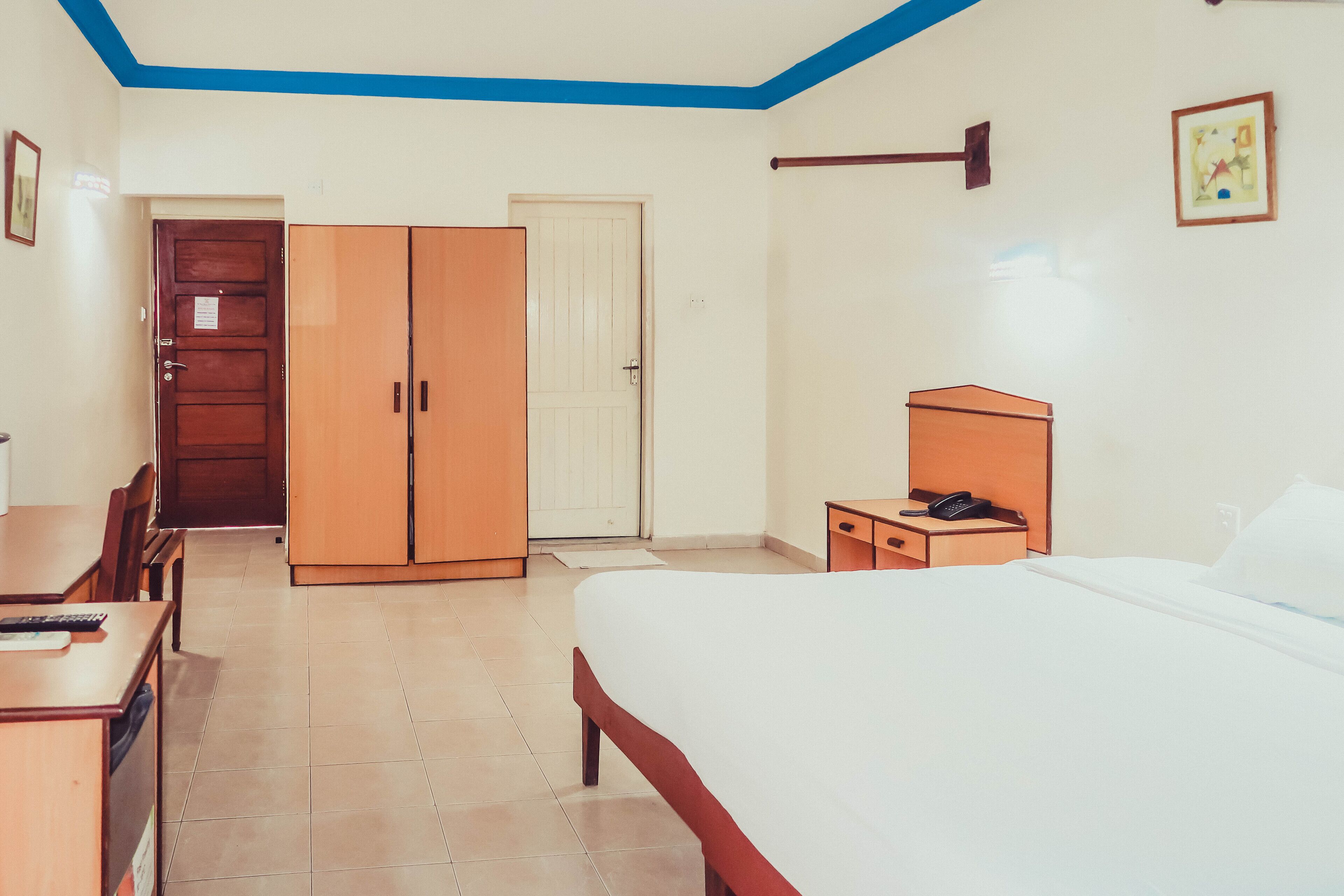 A hotel room with a bed, a desk, a wardrobe, and a bedside table with a phone.