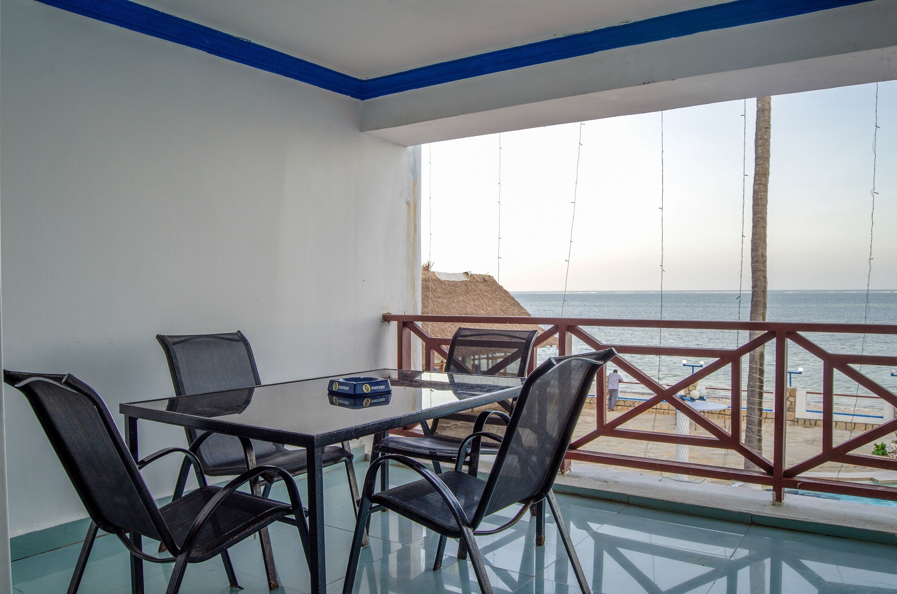 A balcony with a glass table and chairs, overlooking a beach with a wooden fence and a palm tree.