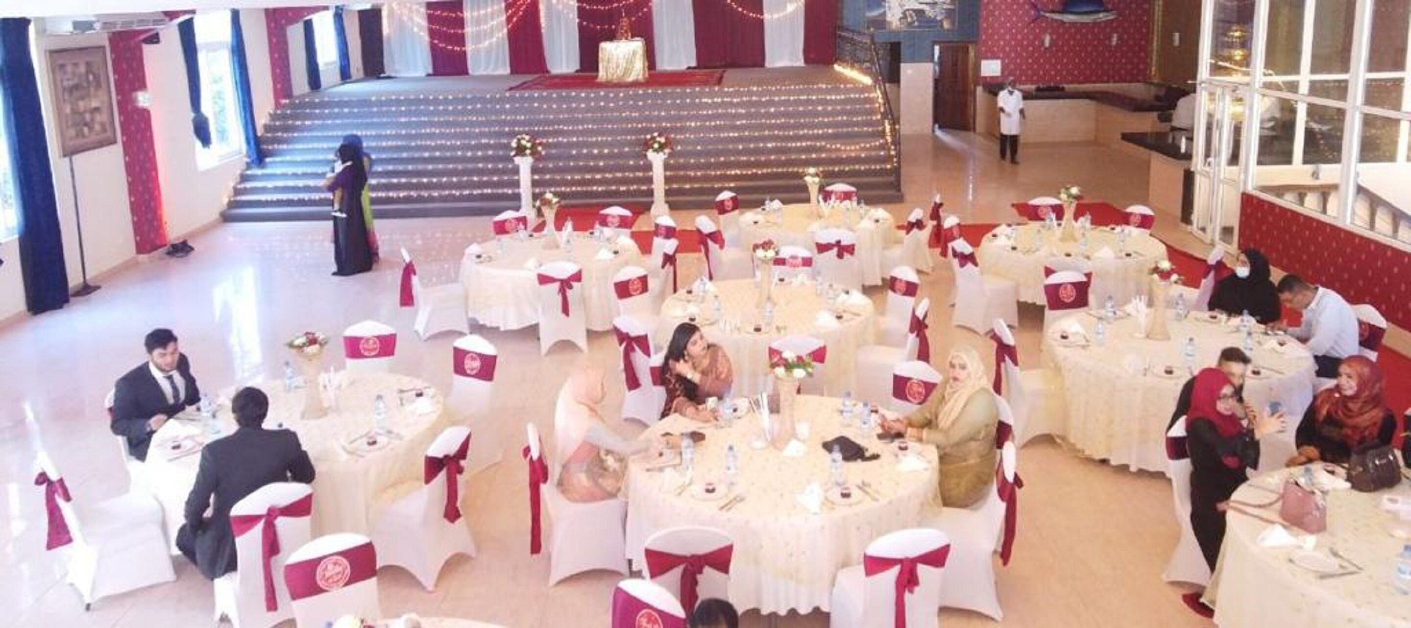 A banquet hall with round tables, white and red decor, and a stage in the background.