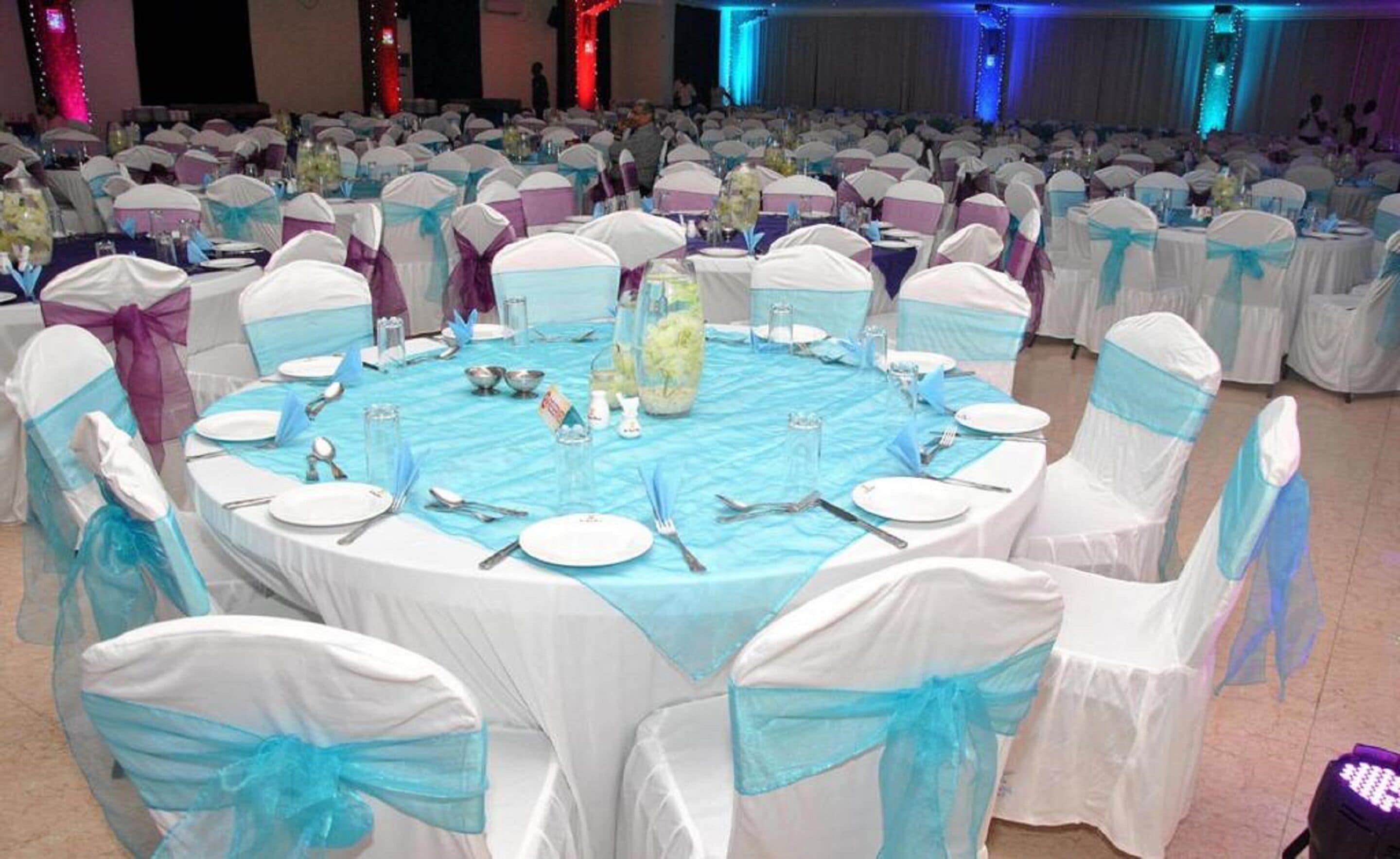A banquet hall with round tables covered in blue tablecloths, surrounded by chairs with white and blue sashes.