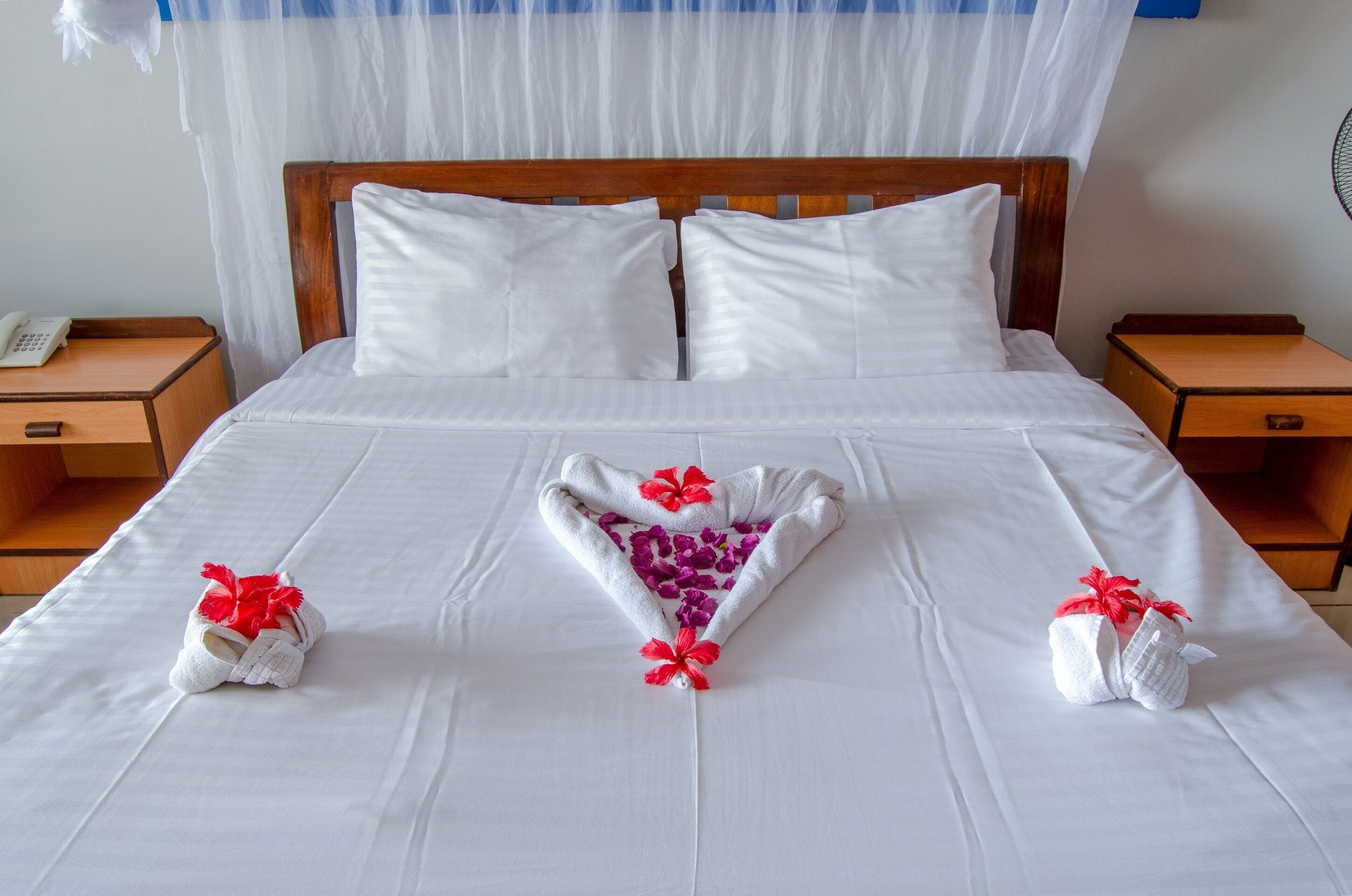 Un lit bien fait, avec du linge de lit blanc et une décoration en forme de cœur réalisée avec des serviettes et des fleurs rouges.