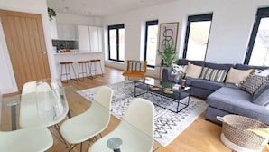 A modern living room with a sofa, coffee table, and chairs. There is a kitchen in the background with white cabinets and a dining area with a glass table and chairs.
