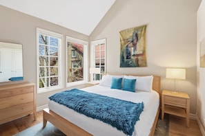 Une chambre à coucher comprenant un lit, une table de chevet, une commode et une fenêtre donnant sur une maison.