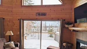 L’intérieur chaleureux d’un chalet avec un plancher en bois, un canapé en cuir, une table basse, une cheminée et une terrasse offrant une vue sur un paysage enneigé.
