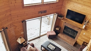 Un intérieur de chalet chaleureux avec une grande fenêtre, une cheminée, un téléviseur et un canapé.